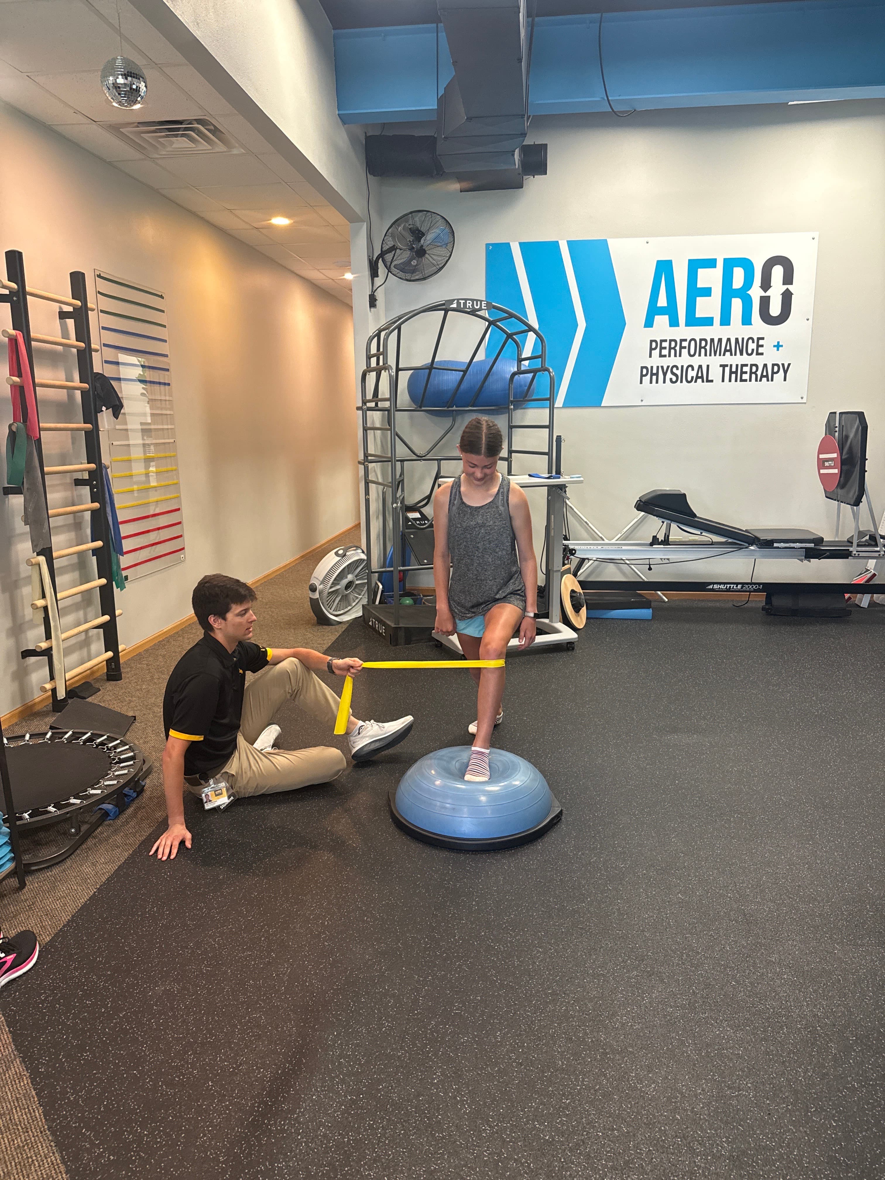 Aero Performance physical therapist guiding a patient through a resistance-band knee stability exercise on a balance dome in the Coralville, Iowa clinic